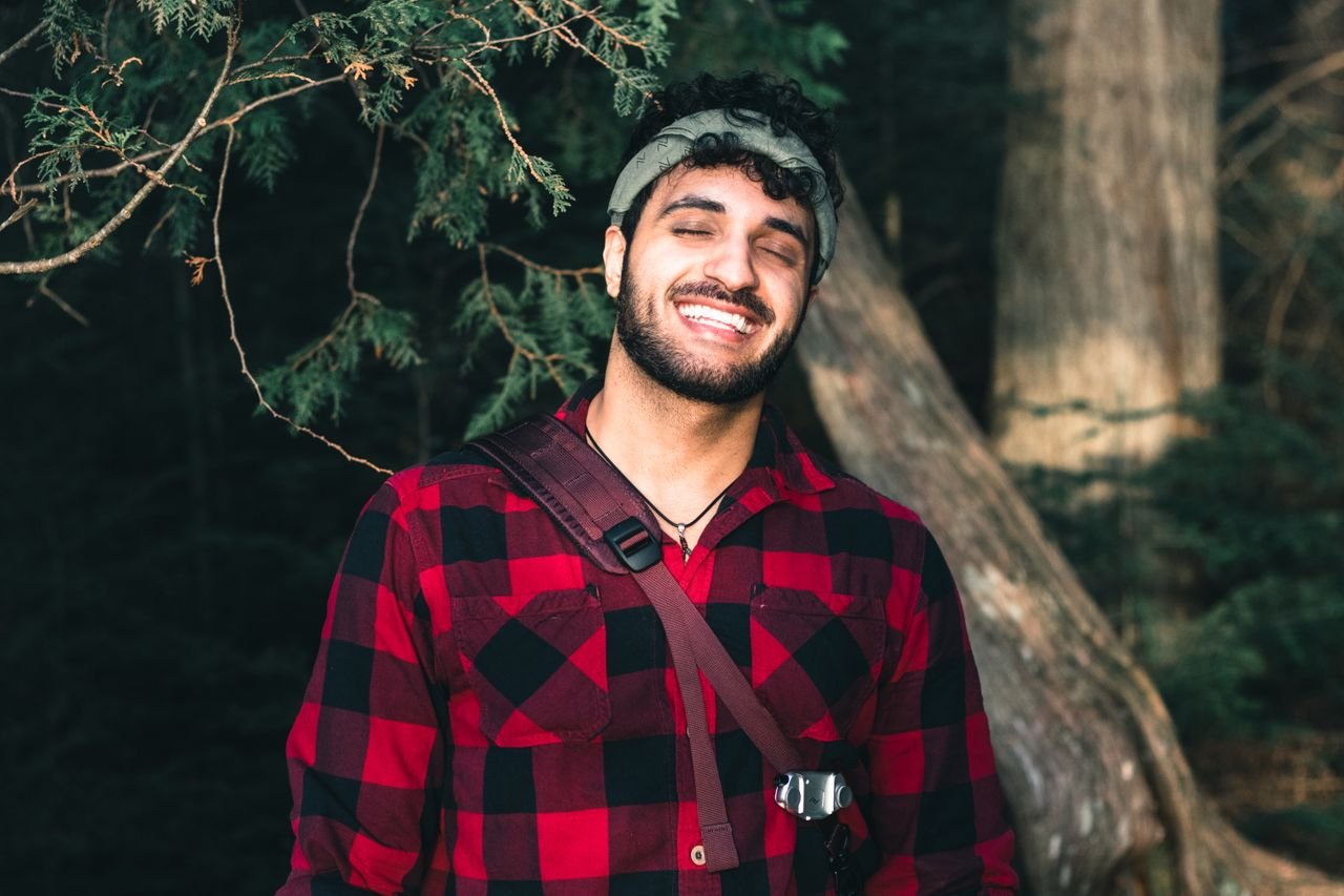 about-us-02 A joyful man in a red plaid shirt smiling in a forest setting.