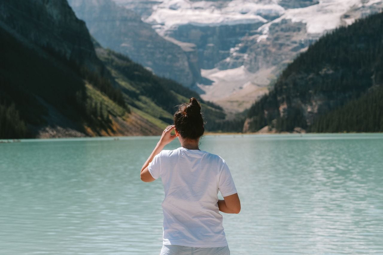 collection-03 A woman in a white t-shirt stands by a mountain lake, enjoying the serene view.